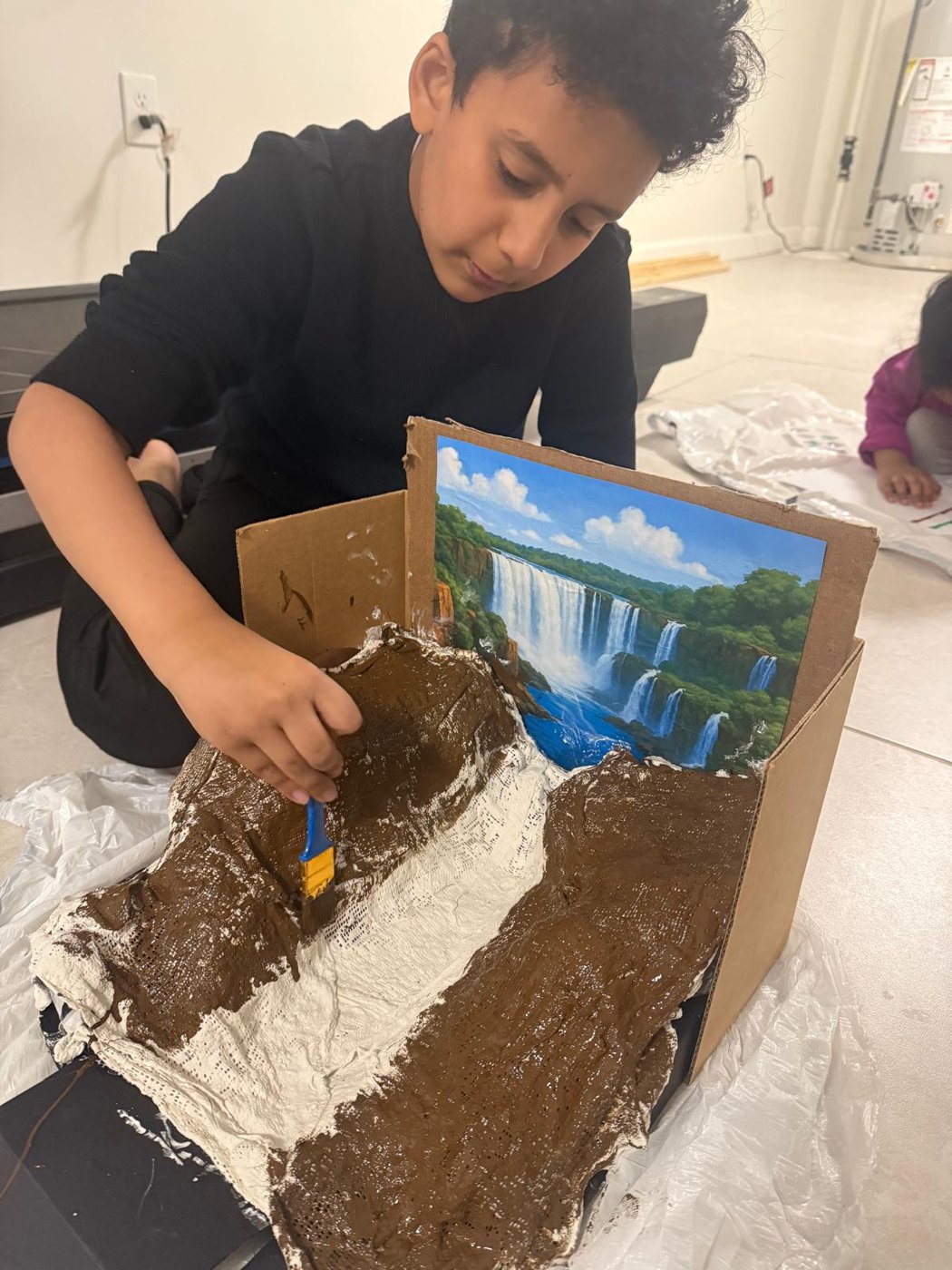 A child painting a landscape model inside a cardboard box with a waterfall backdrop