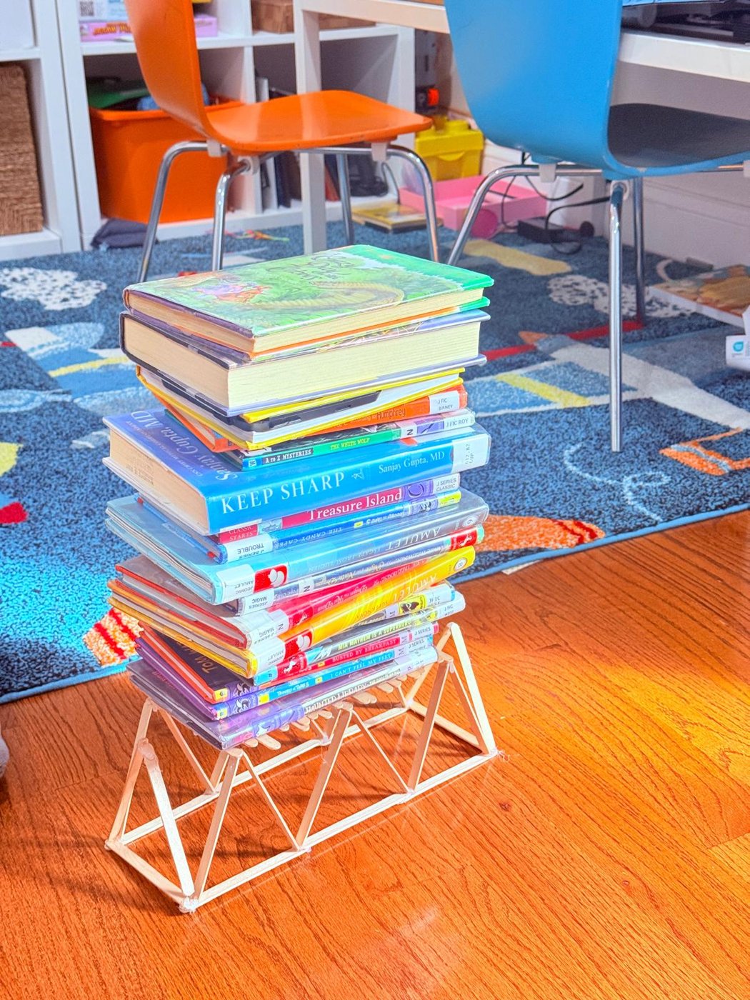 A craft-stick truss bridge holding a tall stack of books on a wooden floor
